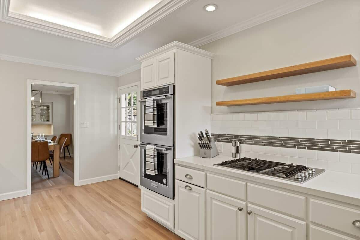 A modern kitchen with white cabinetry, a double oven, a gas stove, and wooden shelves.