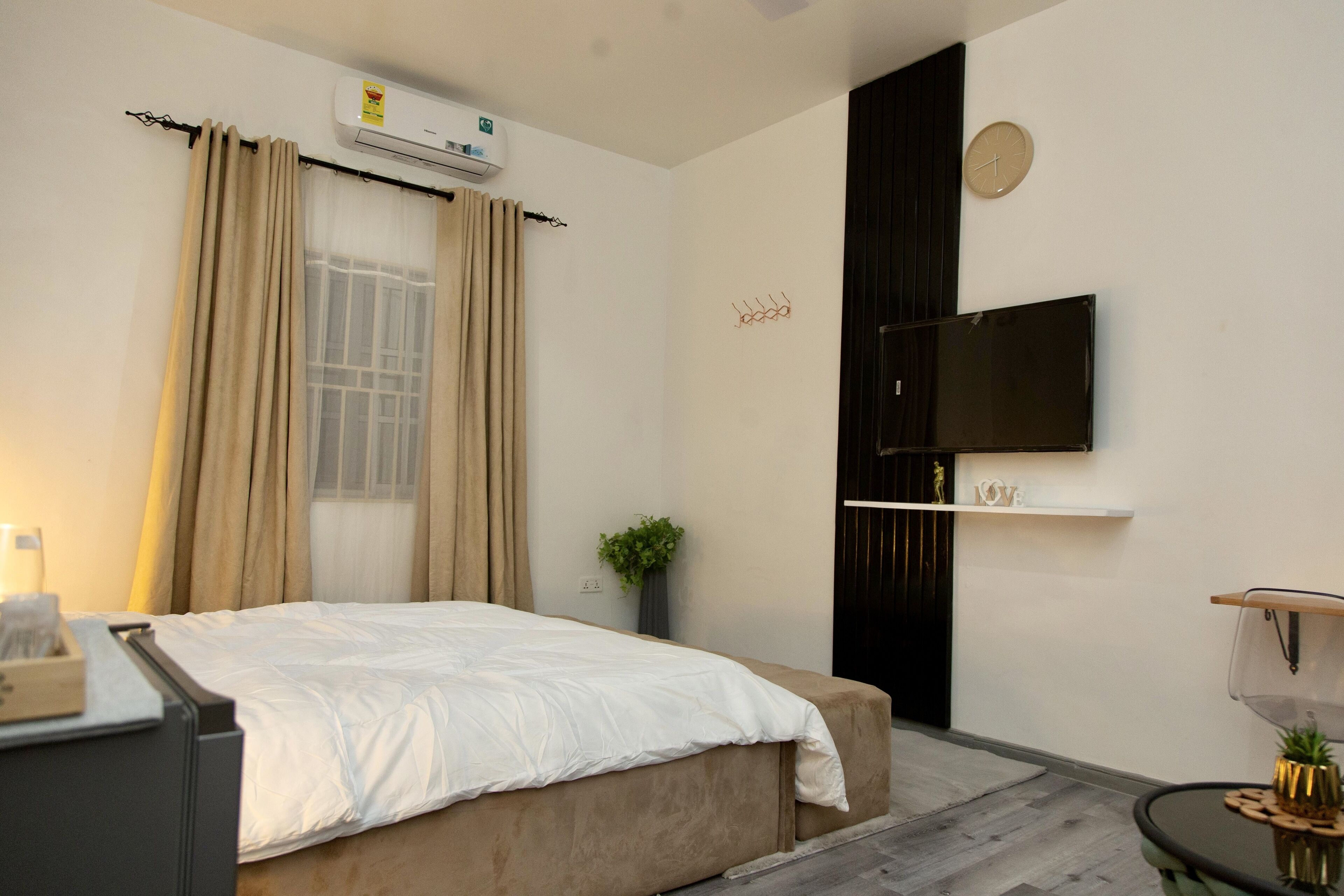 Een slaapkamer met een bed, een aan de muur bevestigde televisie, een nachtkastje en een raam met gordijnen.