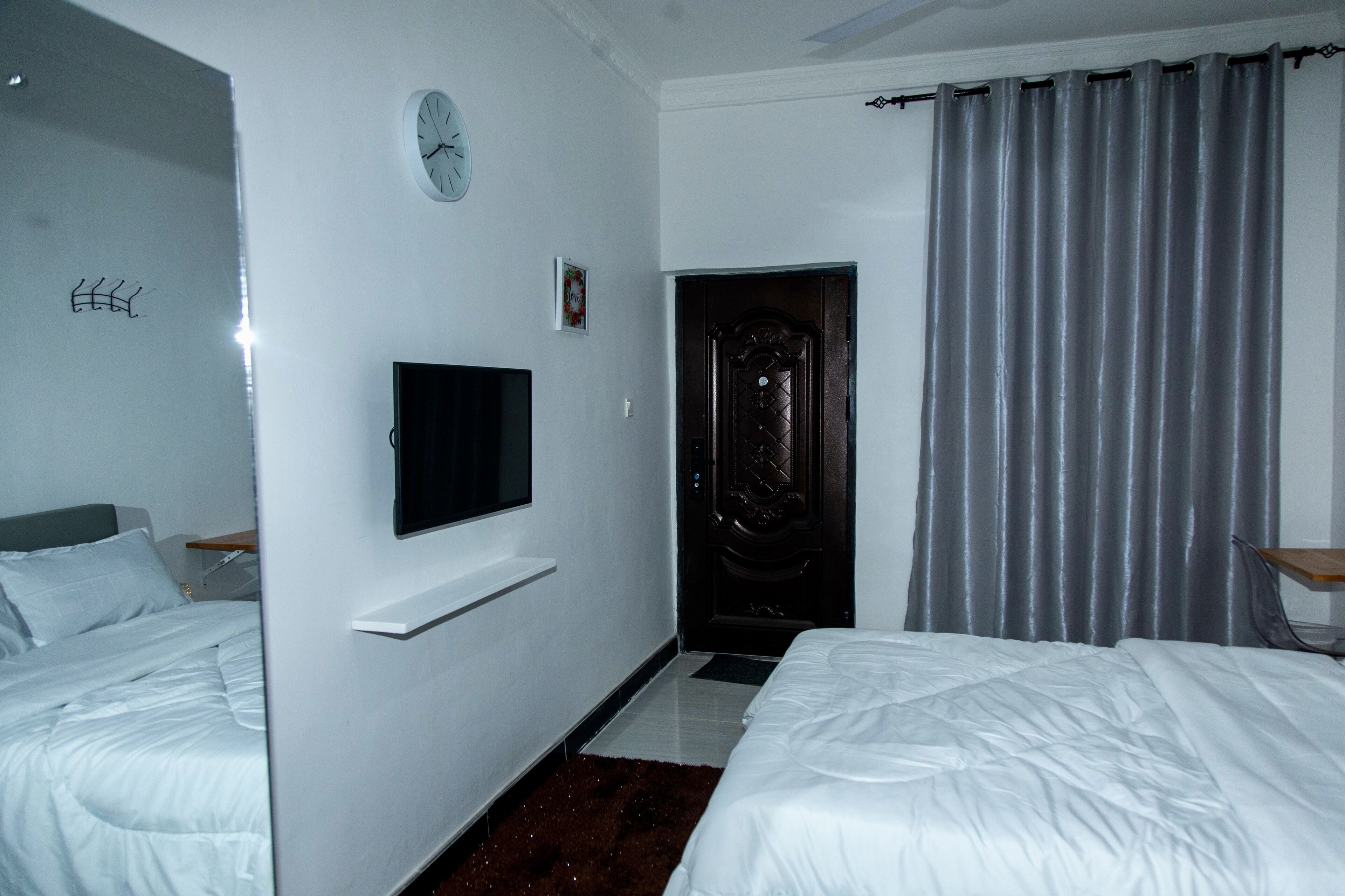 Ein Hotelzimmer mit zwei Betten, einem an der Wand befestigten Fernseher, einer Uhr und einer Tür.