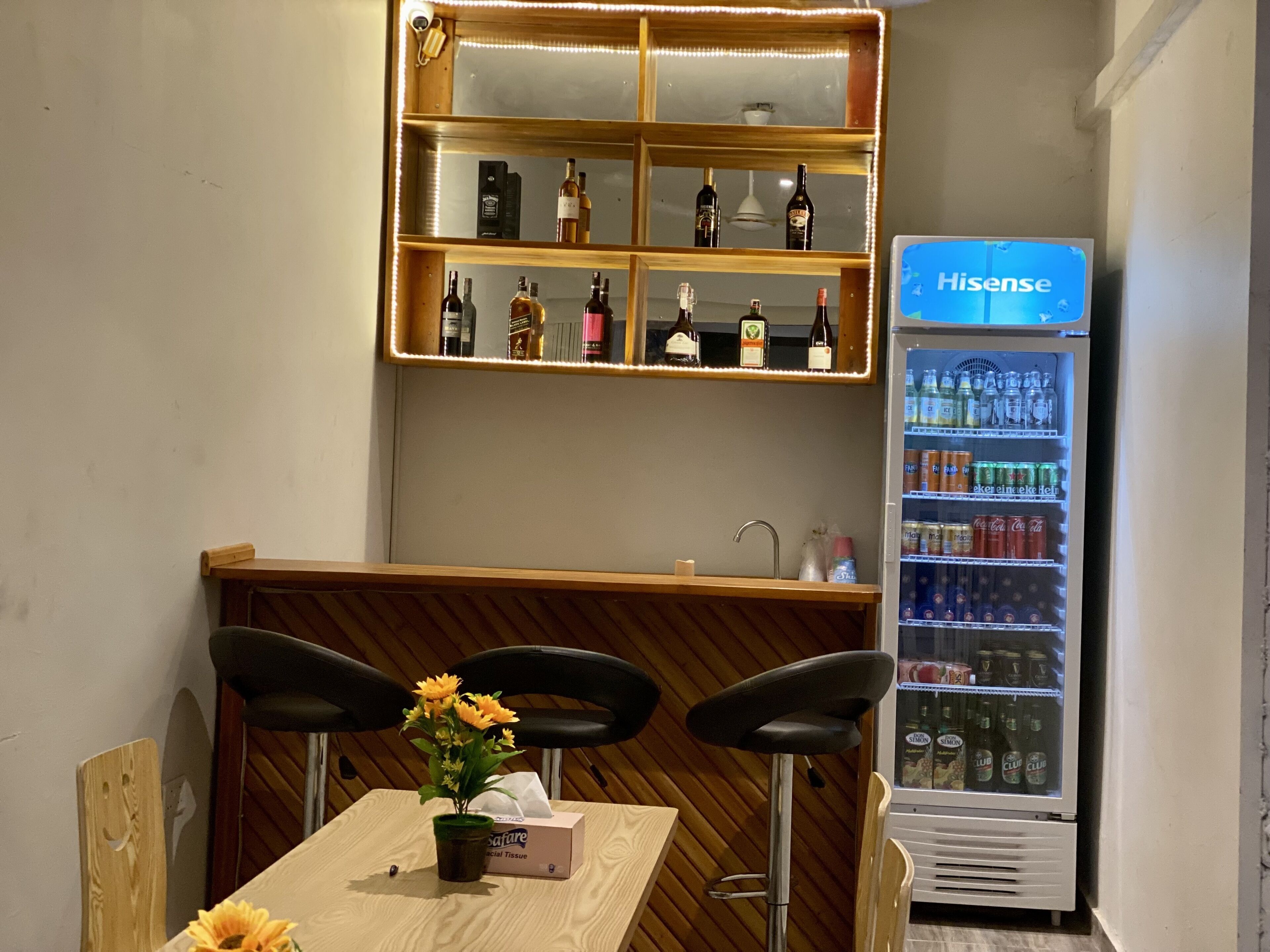 Uma área de bar com balcão de madeira, prateleira com diversos tipos de garrafas e uma geladeira Hisense abastecida com bebidas.