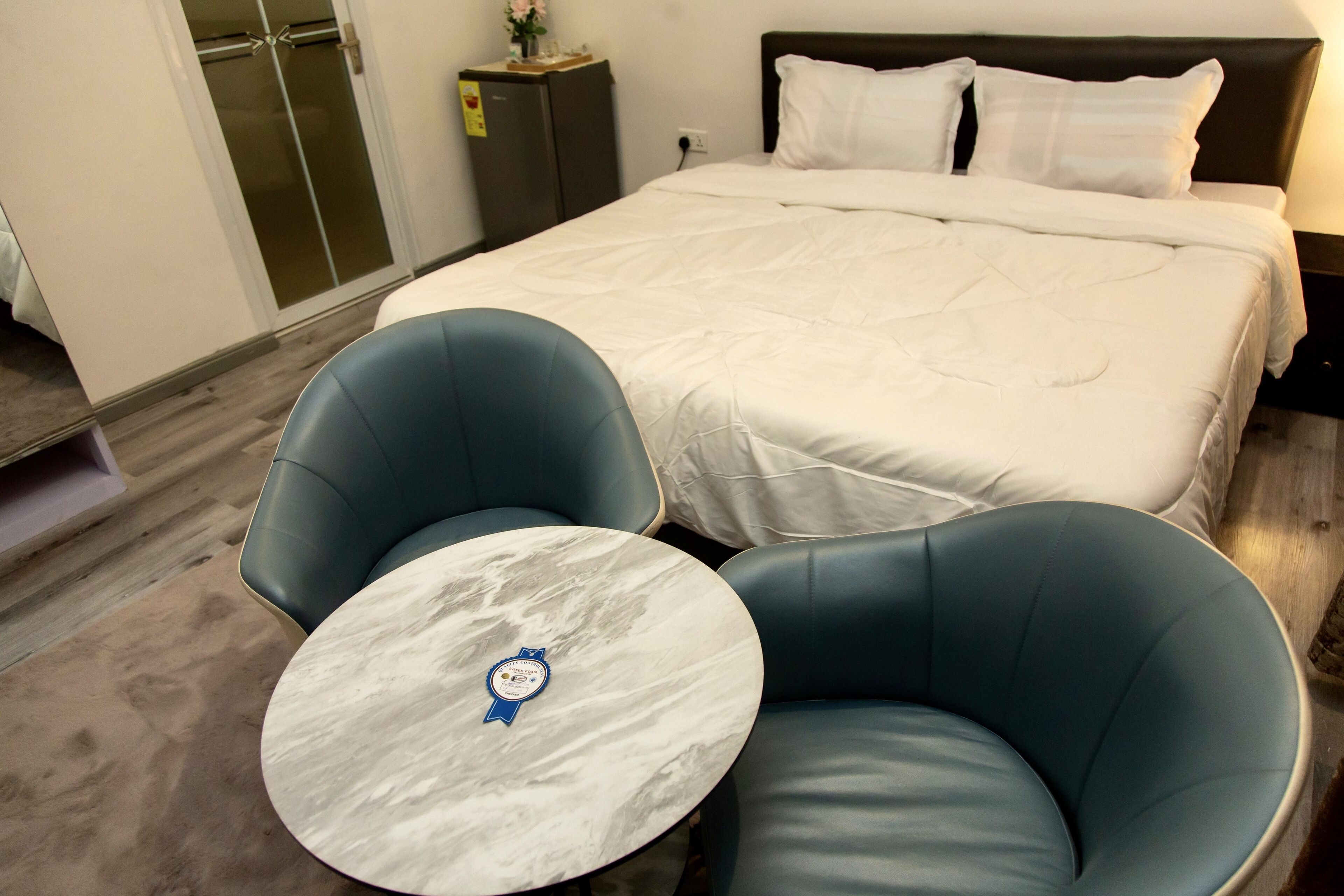 Una camera d'albergo con un letto grande, due poltrone blu, un tavolino in marmo e un comodino con una lampada.