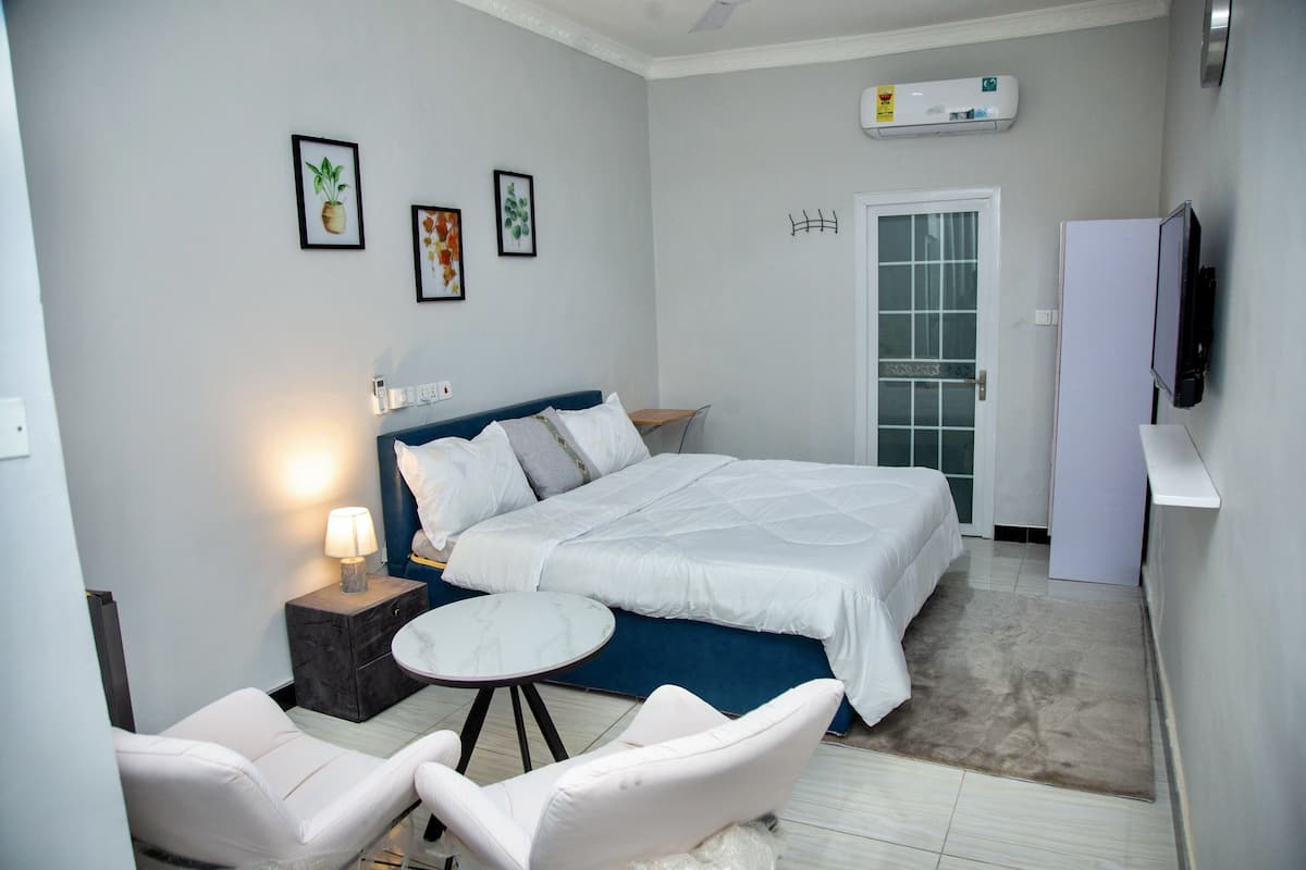 Un dormitorio con una cama, dos sillas blancas, una mesita redonda, una mesita de noche con una lámpara y un televisor en la pared.