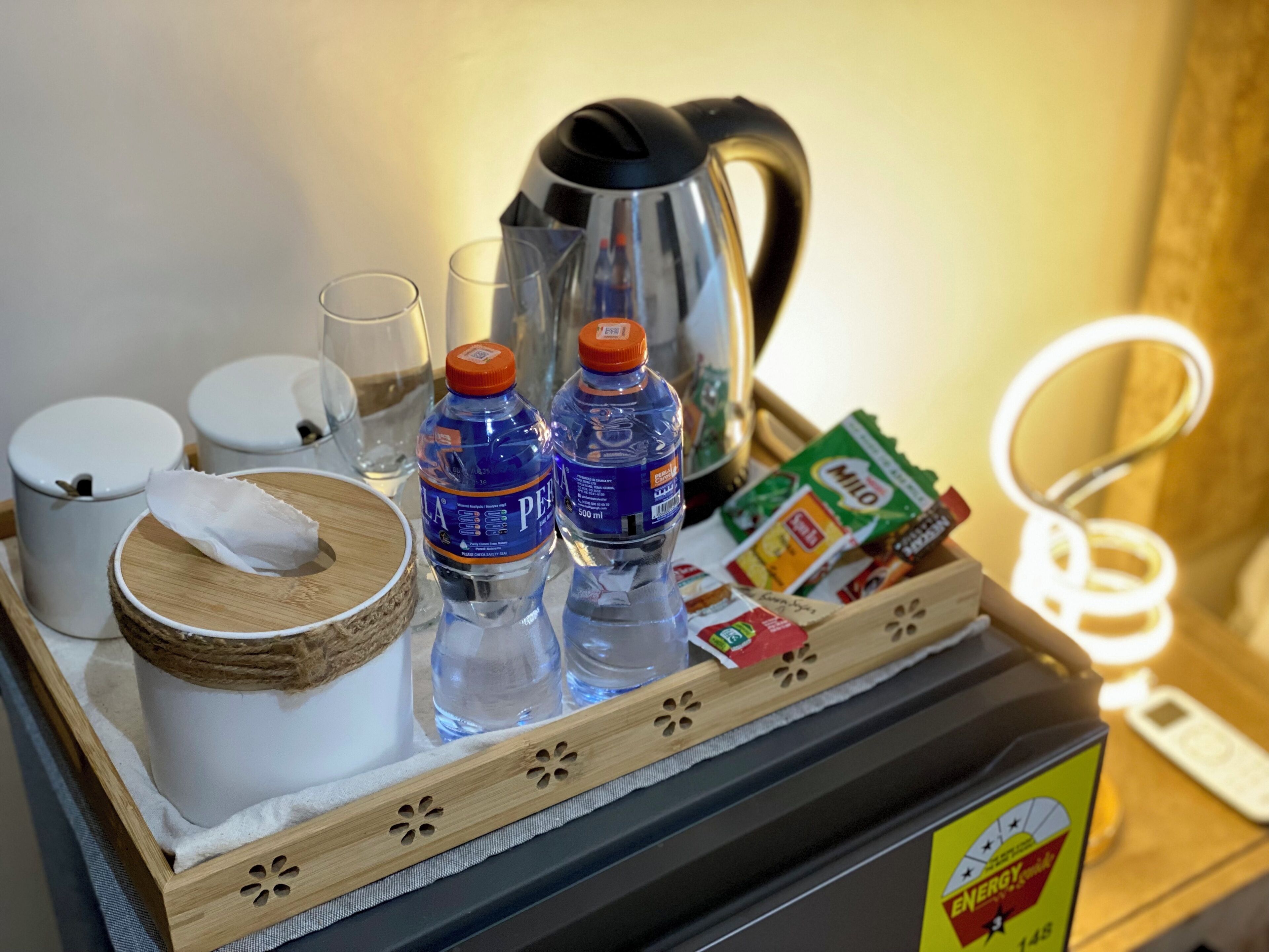 Ein Tablett mit einem Wasserkocher, zwei Wasserflaschen, Taschentüchern, einem Glas und einer Packung Milo-Cereal.