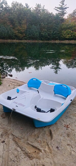 Un pequeño barco de plataforma blanco y azul con la etiqueta 'sun slider', amarrado en una orilla arenosa junto a un lago tranquilo con árboles al fondo.