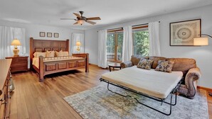 Ein Schlafzimmer mit einem hölzernen Bett, einer Schlafcouch, einem Deckenventilator und einem großen Fenster mit Vorhängen.