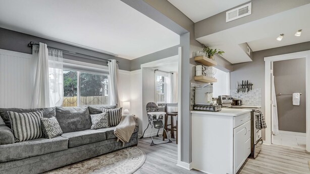 Een moderne woonkamer met een grijze bank, een rond vloerkleed en een keuken met witte kasten en een klein eiland.