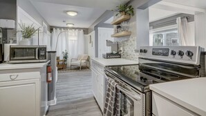Een moderne keuken met witte kastjes, een inbouwmagnetron, een fornuis en een spoelbak. Boven het fornuis staat een plant op een plank en er staat een stoel op de achtergrond.