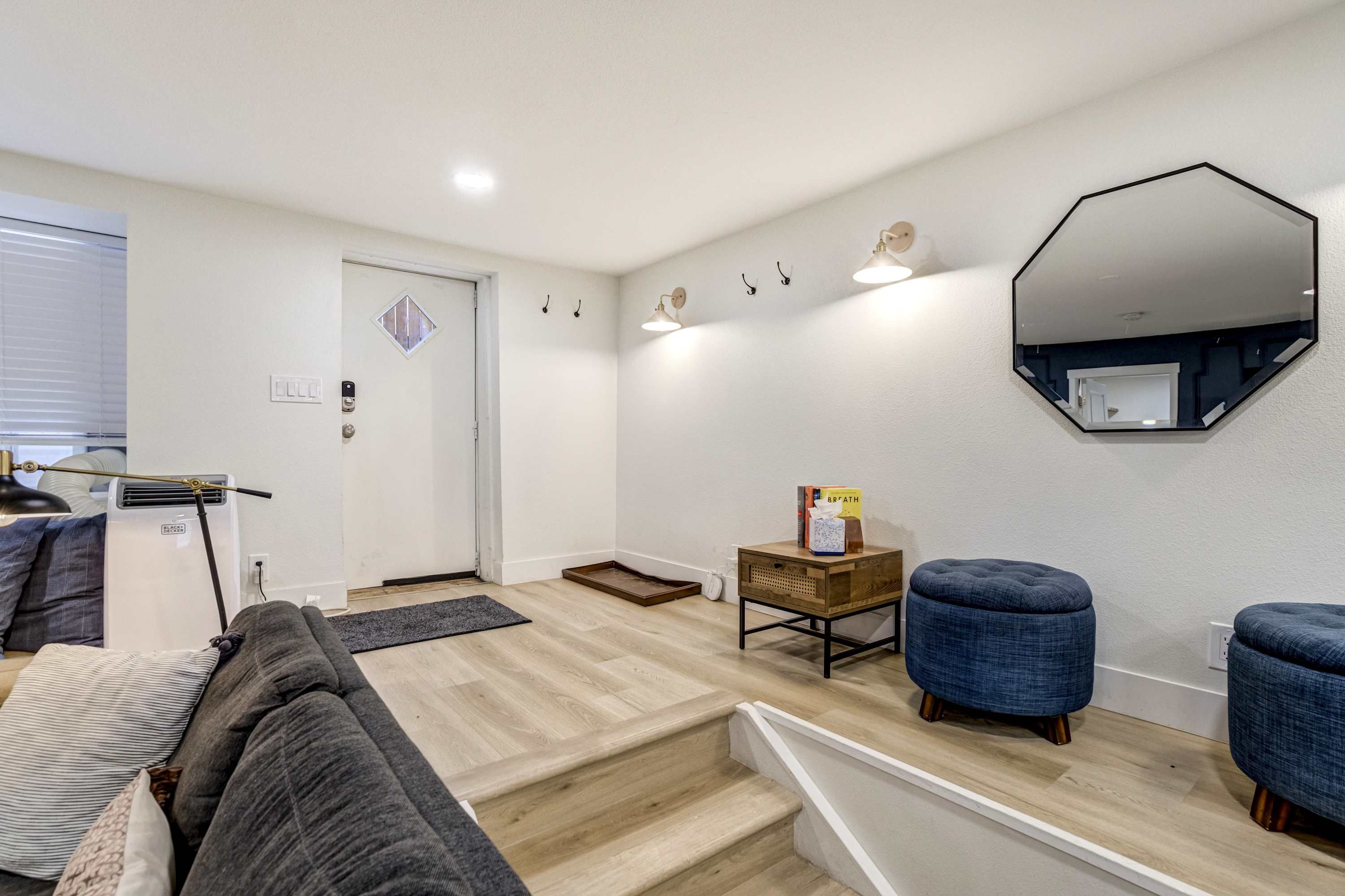En moderne stue med sofa, skammel og sofabord. Der er en dør med et vindue, et spejl på væggen og et lille træsidebord med en bog og vatrondeller.