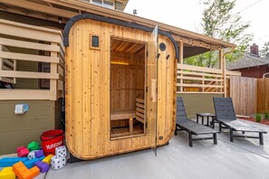 A sauna with a wooden exterior, a bench inside, and two lounge chairs outside.