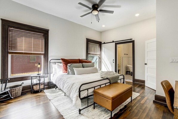 A bedroom with a bed, bedside tables, a bench, a ceiling fan, and a view of the outside.