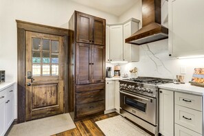 Una cocina con puerta de madera, armarios blancos, estufa y cafetera.