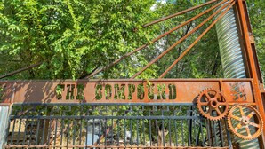 A rusty metal gate with the words 'THE COMPOUND' and two large gears, surrounded by greenery.