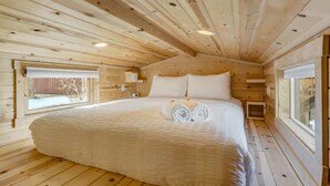 A wooden cabin bedroom with a large bed, two windows, and a sloped ceiling.