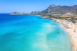 A coastal landscape with clear turquoise waters, a sandy beach, and rocky mountains in the background.