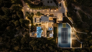 An aerial view of a modern house with a swimming pool, tennis court, and landscaped garden.