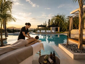 Un espacio de spa con una mesa de masaje, una piscina, sillas de descanso y un edificio pequeño al fondo.