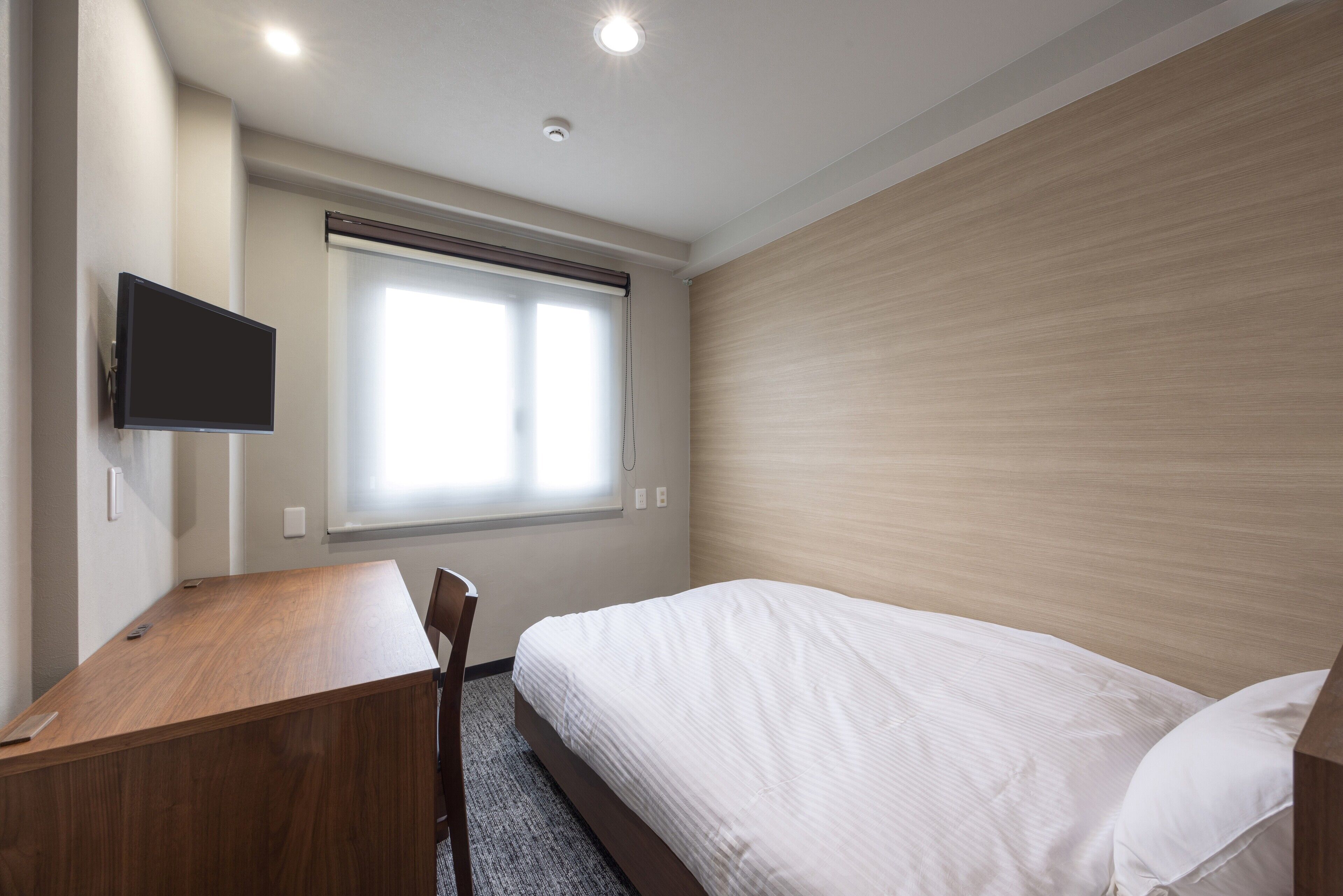 A hotel room with a wooden desk, a bed with white bedding, a mounted TV, and a window with blinds.