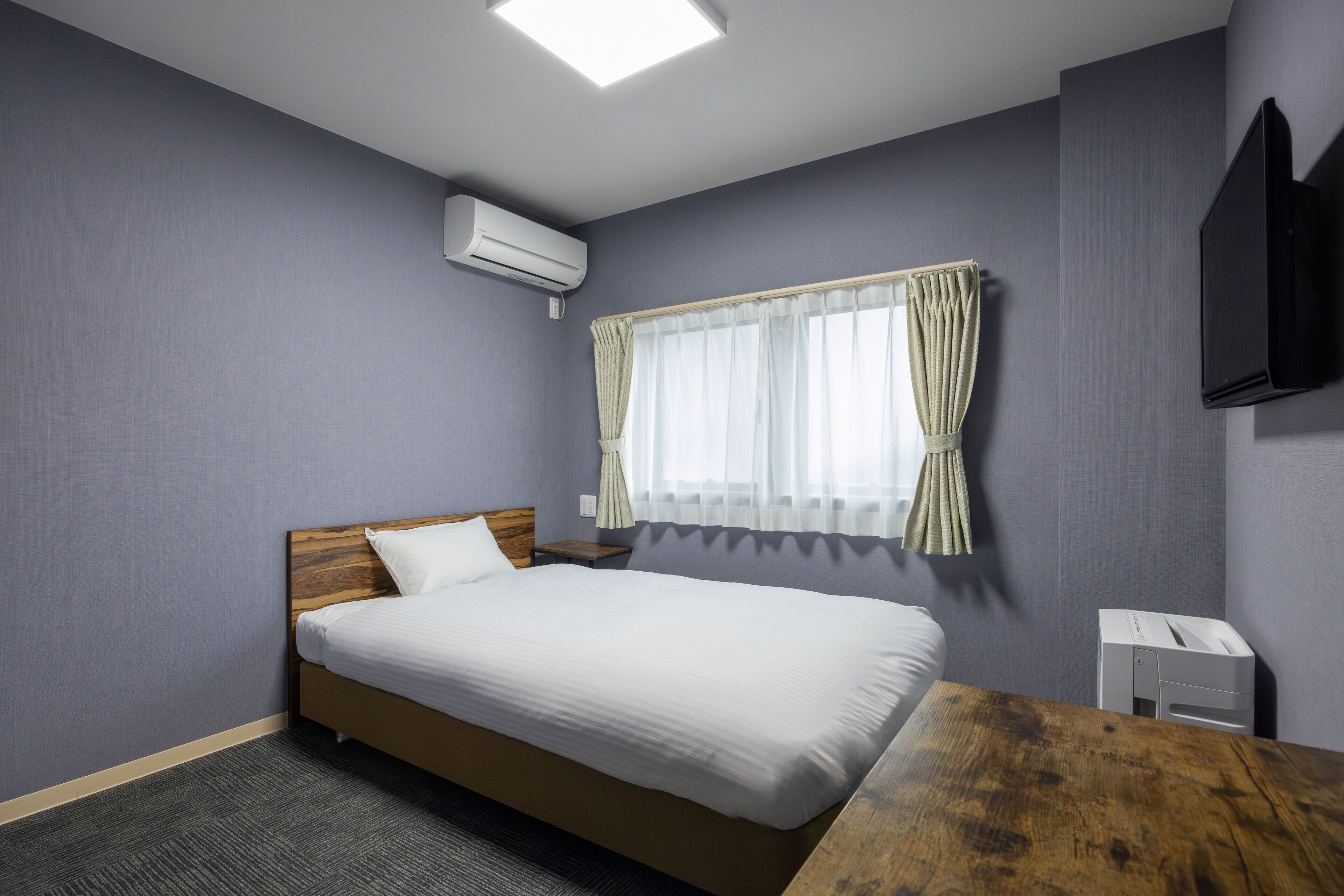 Habitación de hotel con una cama, una mesita de noche de madera, un escritorio, un televisor de pantalla plana, un equipo de aire acondicionado y una ventana con cortinas.