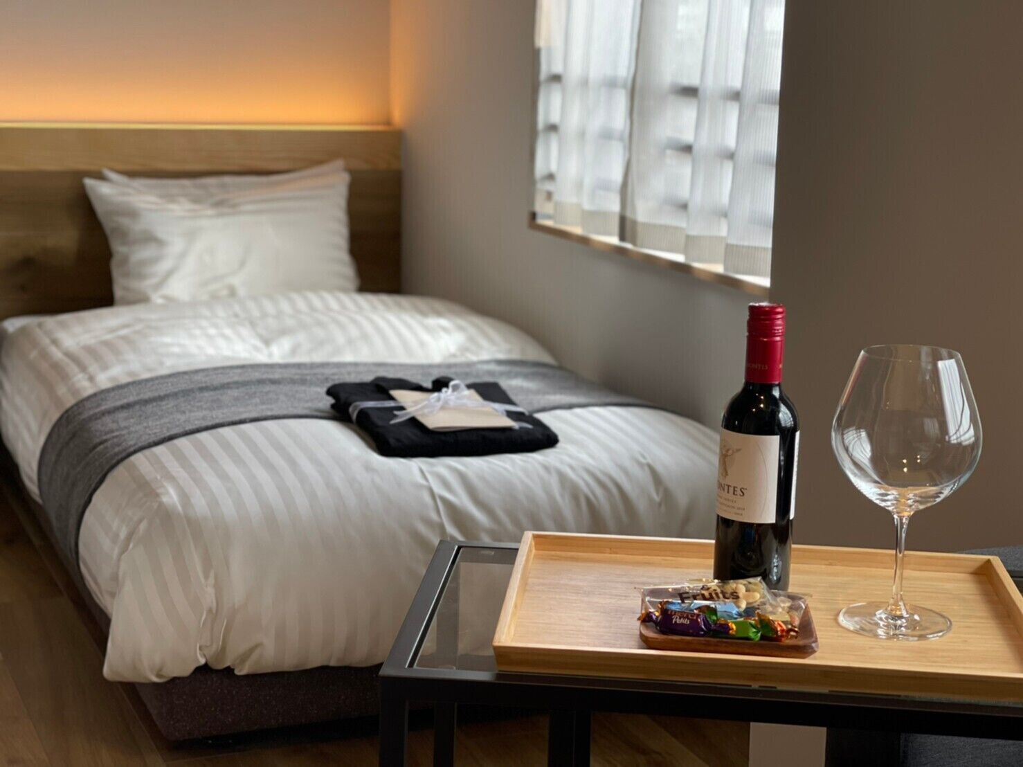 Un lit bien fait, avec une bouteille de vin et un verre sur un plateau, une petite table avec des fruits et une fenêtre avec des stores.