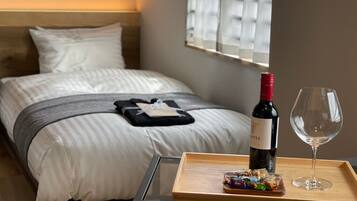 A neatly made bed with a wine bottle and glass on a tray, a small table with fruits, and a window with blinds.