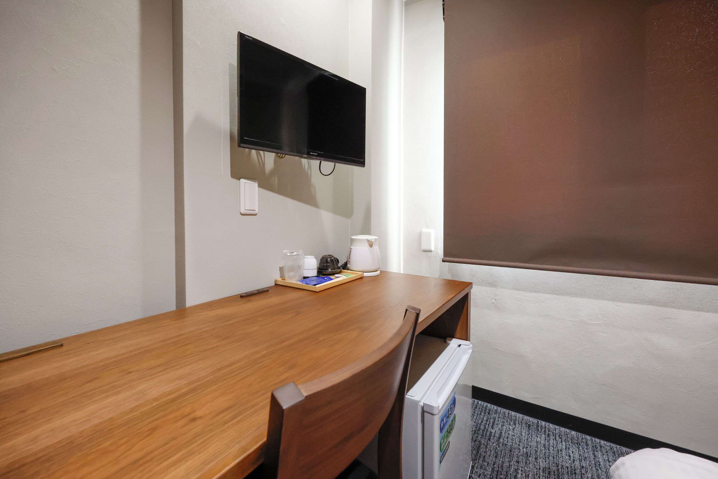 Habitación de hotel con escritorio de madera, televisor de pantalla plana en la pared, pequeña mesa con tetera y tazas, y ventana con persiana marrón.