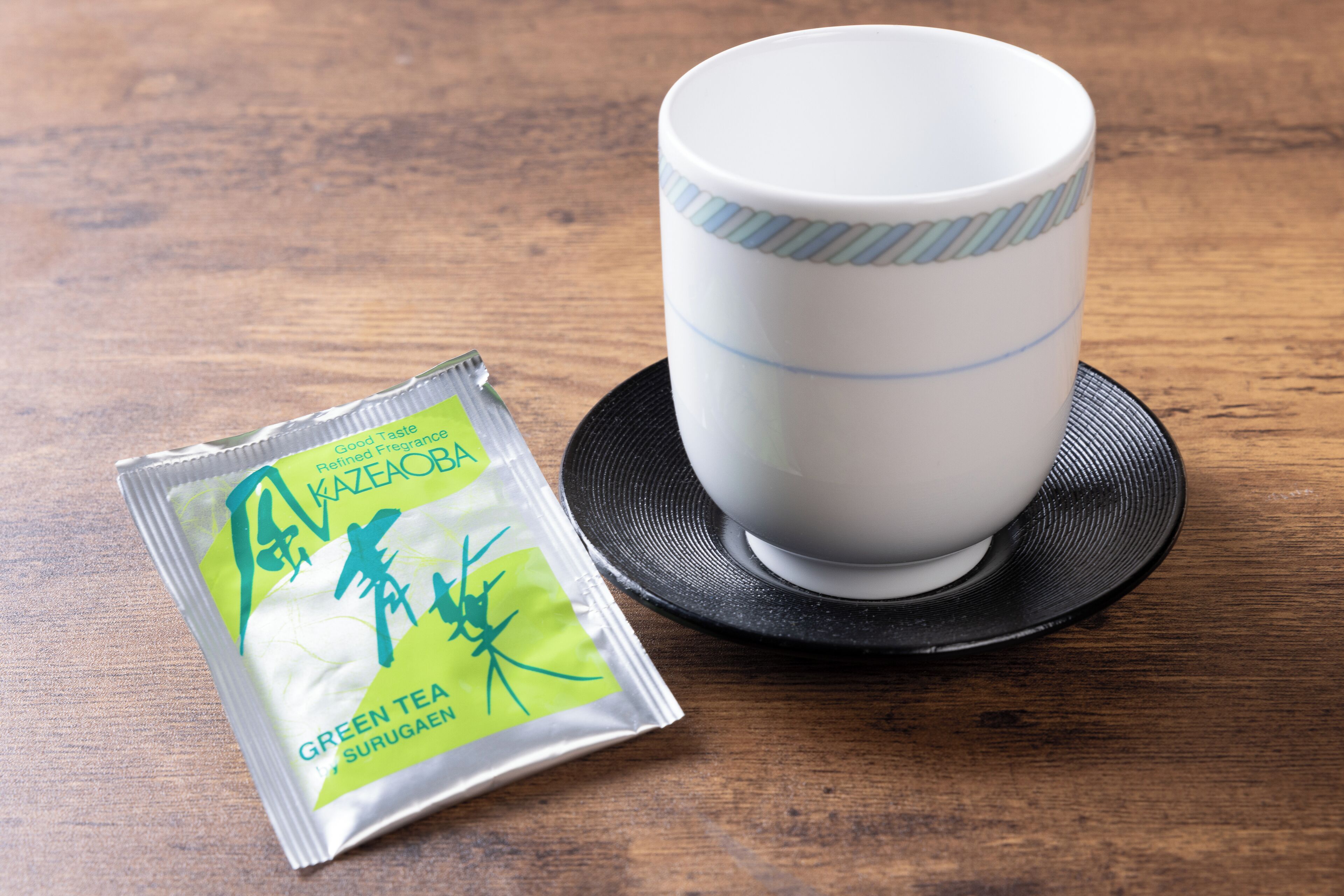 A white cup with a striped pattern and a black saucer on a wooden table. Next to it is a packet of green tea labeled "KAZEOKA" and "GREEN TEA SURUGAEN".