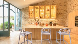 A kitchen with a stone wall, wooden cabinets, and a large window. There are three white bar stools and a countertop with various items. - La Glycine (Tonnac)