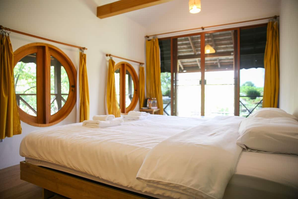 Een net opgemaakt bed met witte lakens en een houten hoofdbord in een kamer met grote ramen en gele gordijnen.