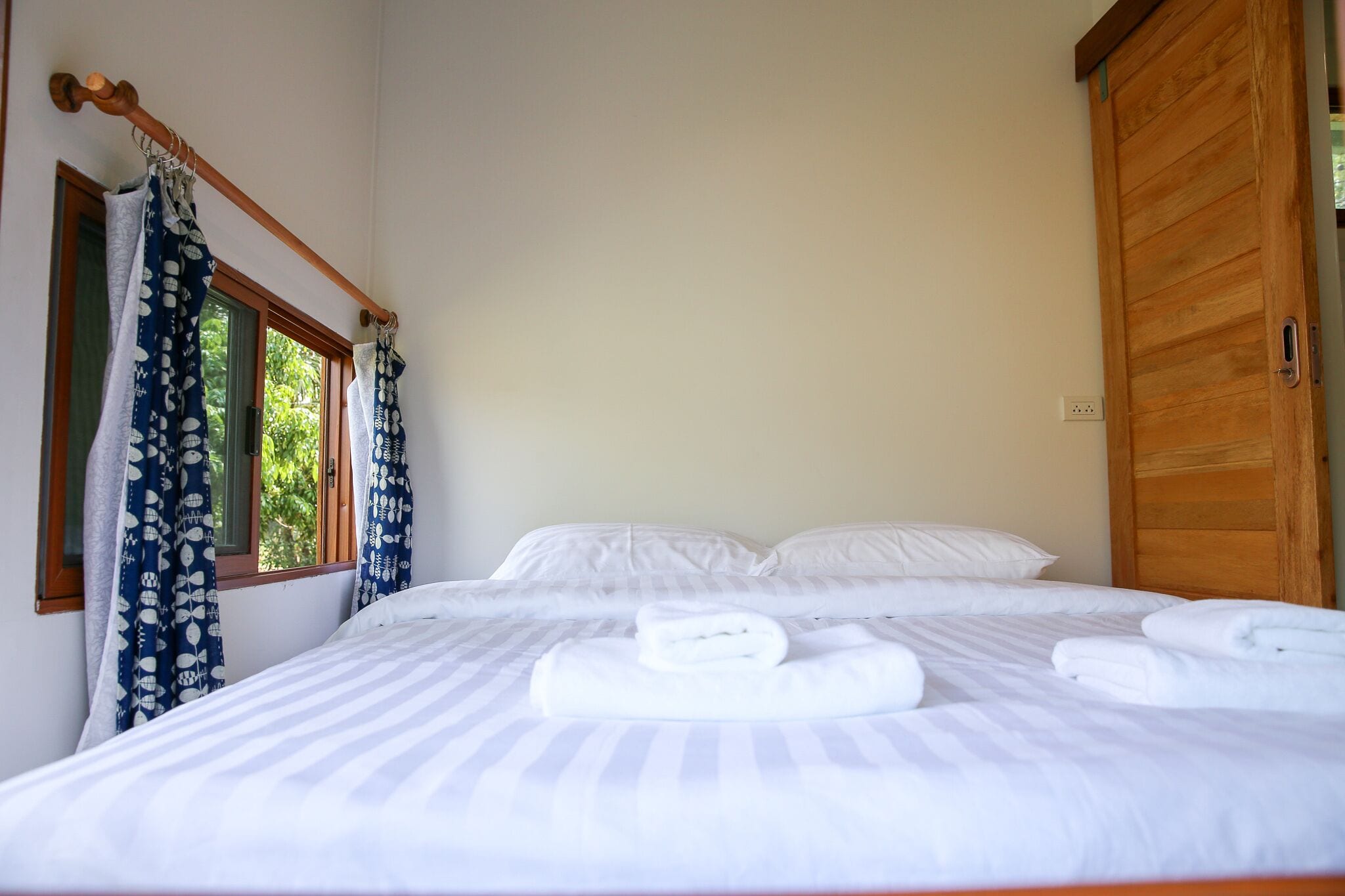 A neatly made bed with white linens and towels, a wooden wardrobe, and a window with curtains.