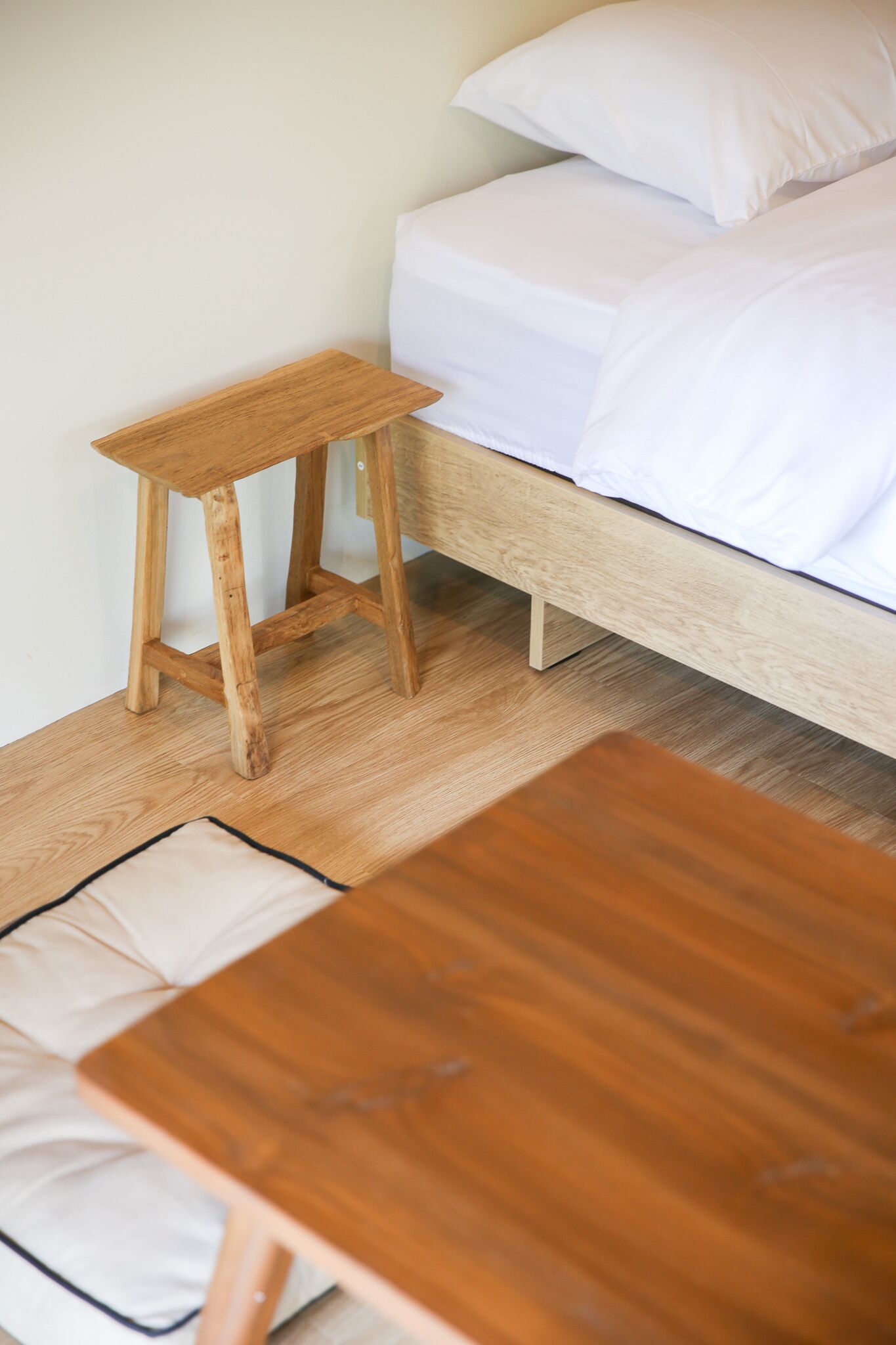 Een houten nachtkastje en een klein houten krukje naast een bed met witte beddengoed.