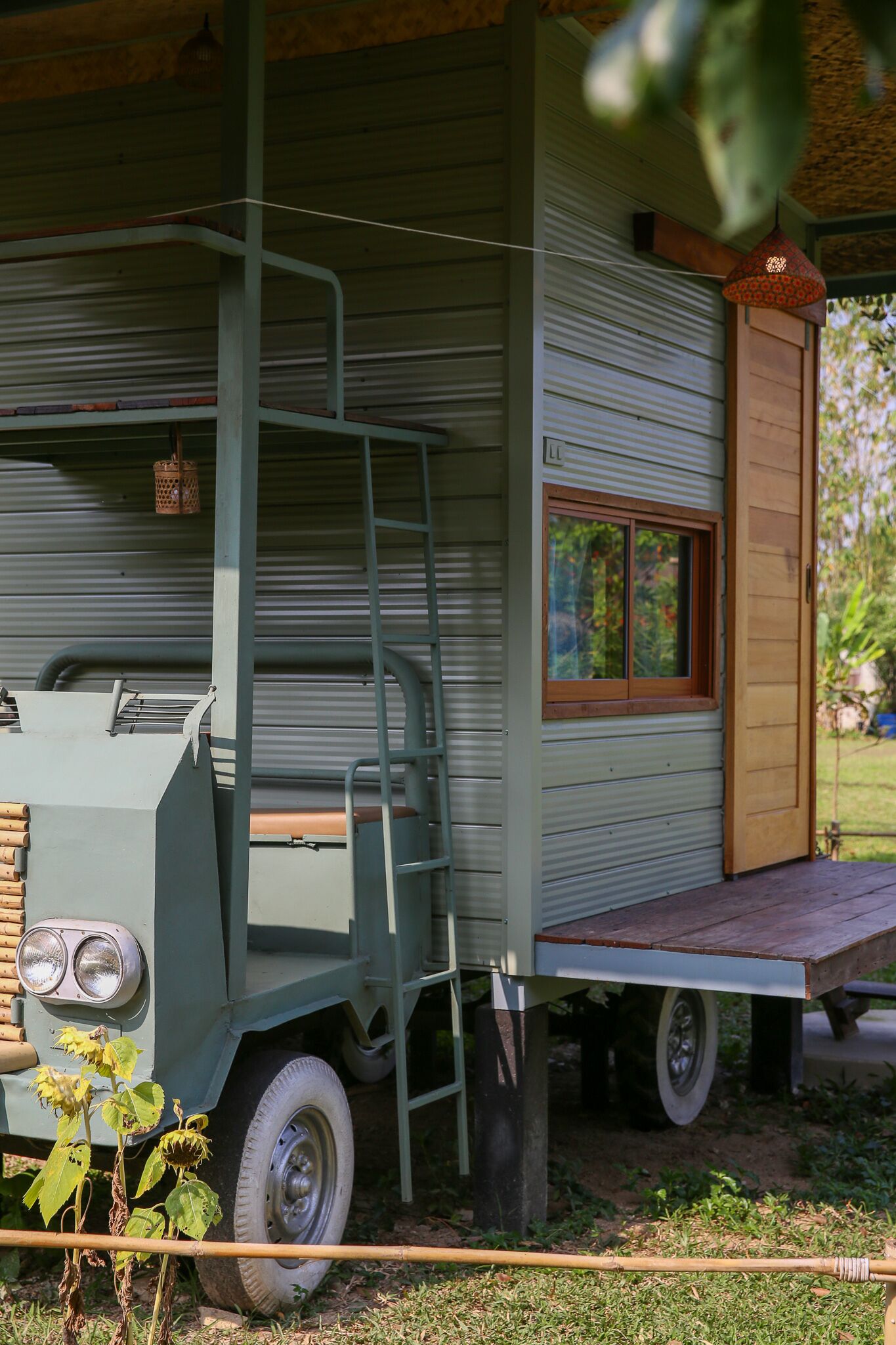 Une maison mobile verte avec une terrasse en bois, une échelle et une fenêtre donnant sur un paysage verdoyant.