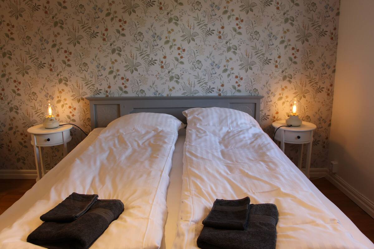 A double bed with white linens and two dark towels folded at the foot.
