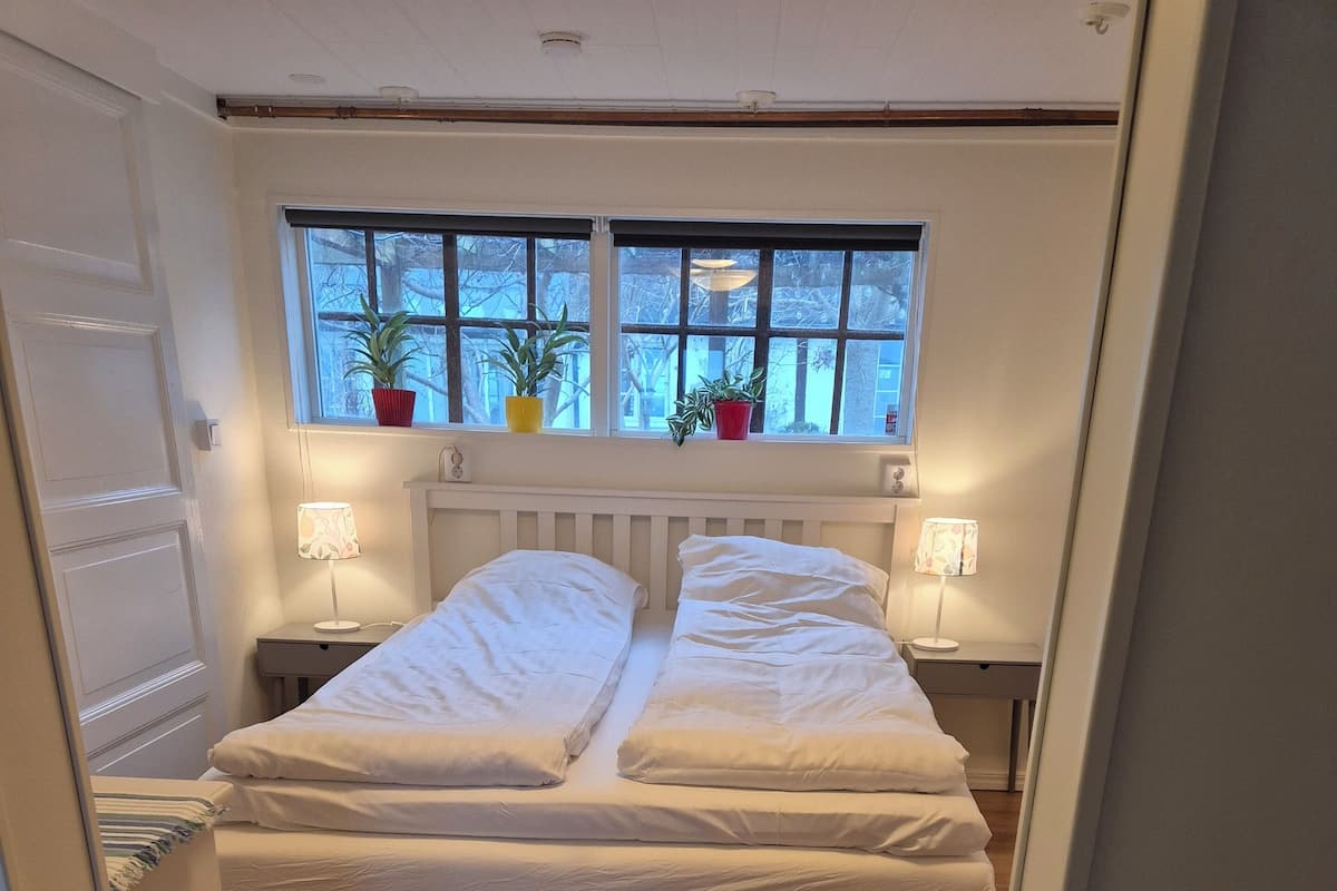 A bedroom with two beds, wooden flooring, a window with plants, and bedside lamps.