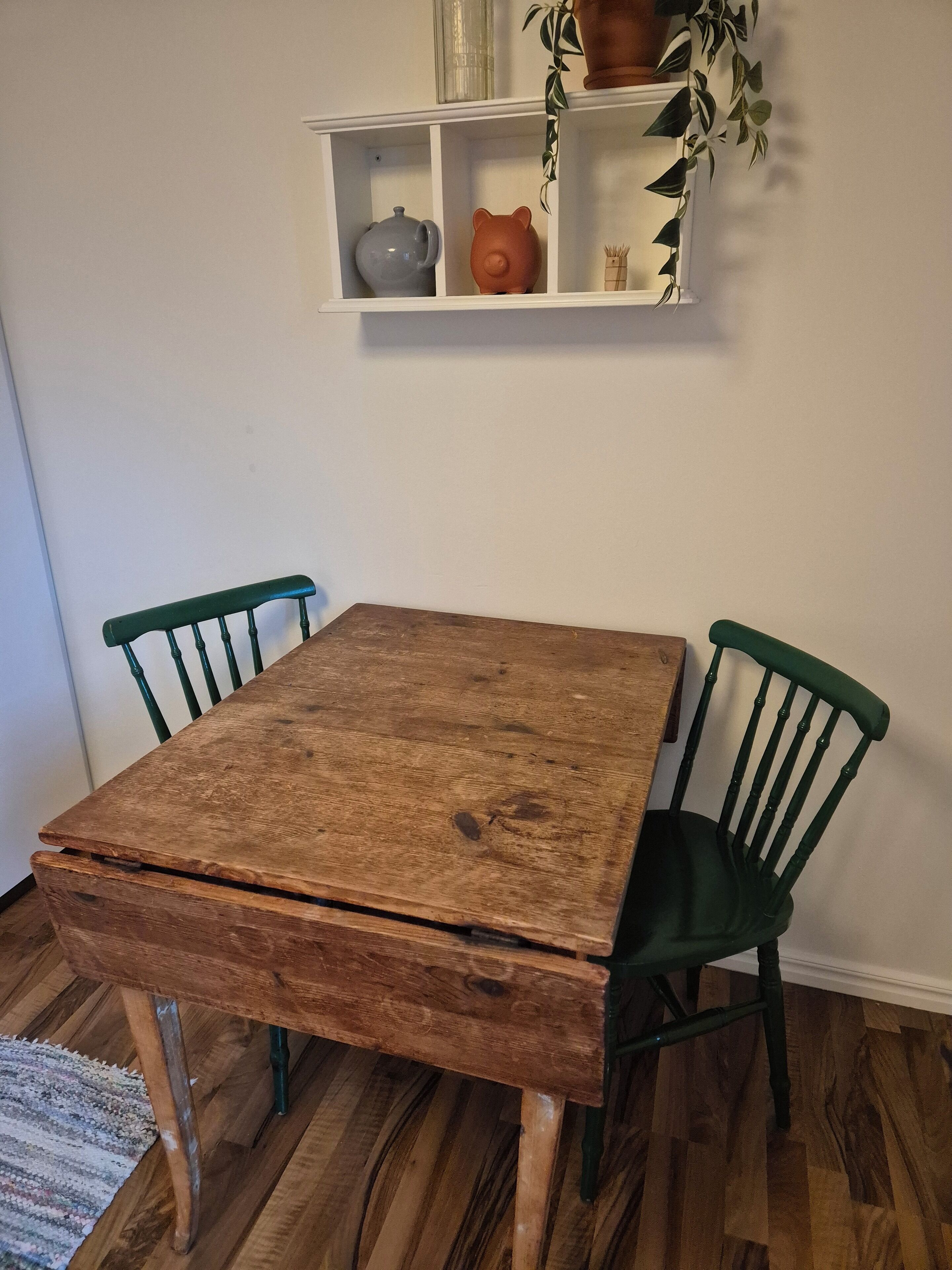 A rustic wooden table with green chairs, a white shelf with decorative items, and a wooden floor.