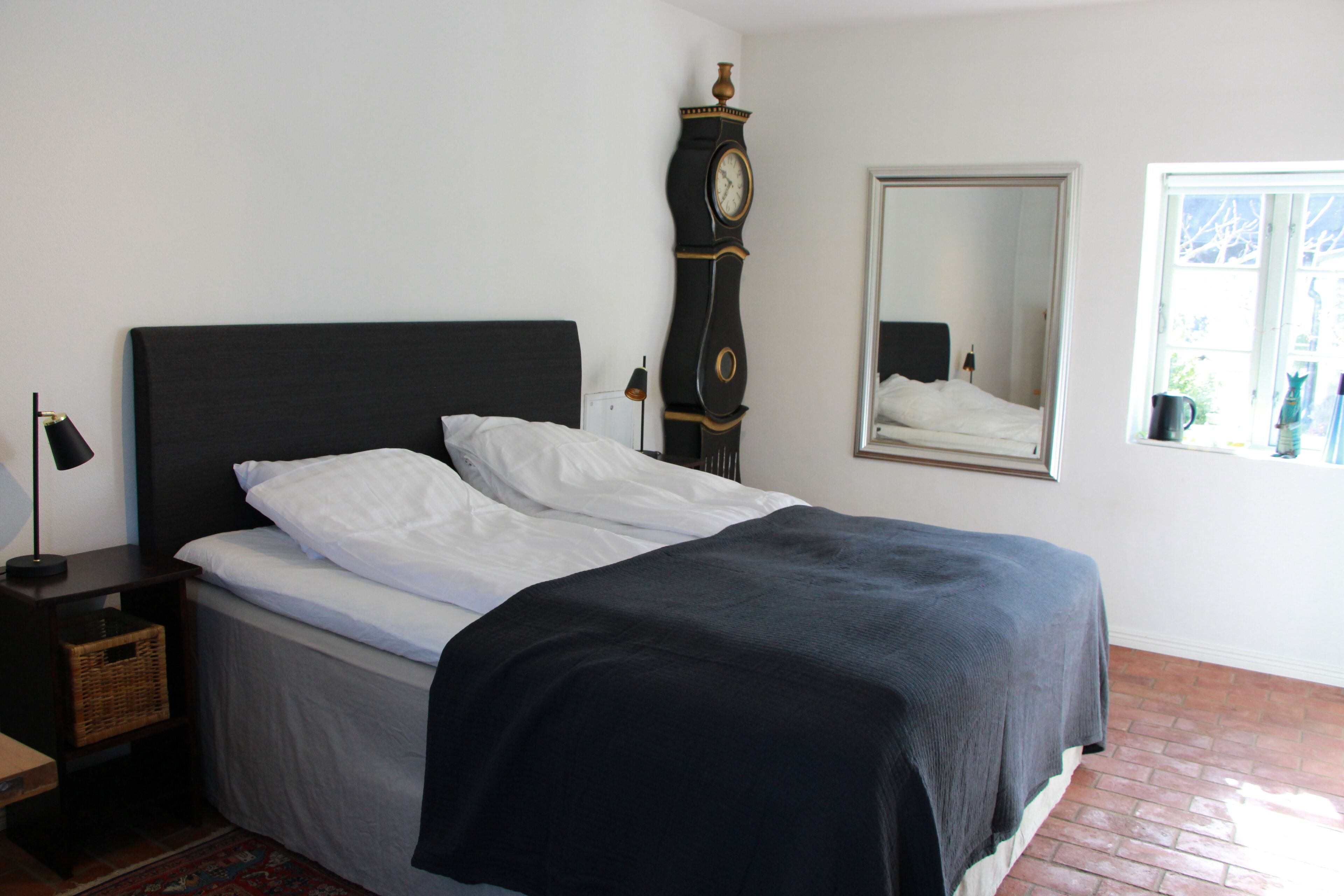 A neatly made bed with white and dark bedding, a tall wall clock, a mirror, and a window with a view.
