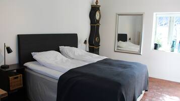 A neatly made bed with white and dark bedding, a tall wall clock, a mirror, and a window with a view.