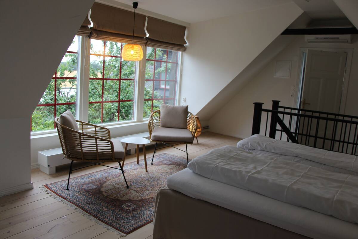 A bedroom with a large bed, a window with a view of greenery, a wicker chair, and a small table.
