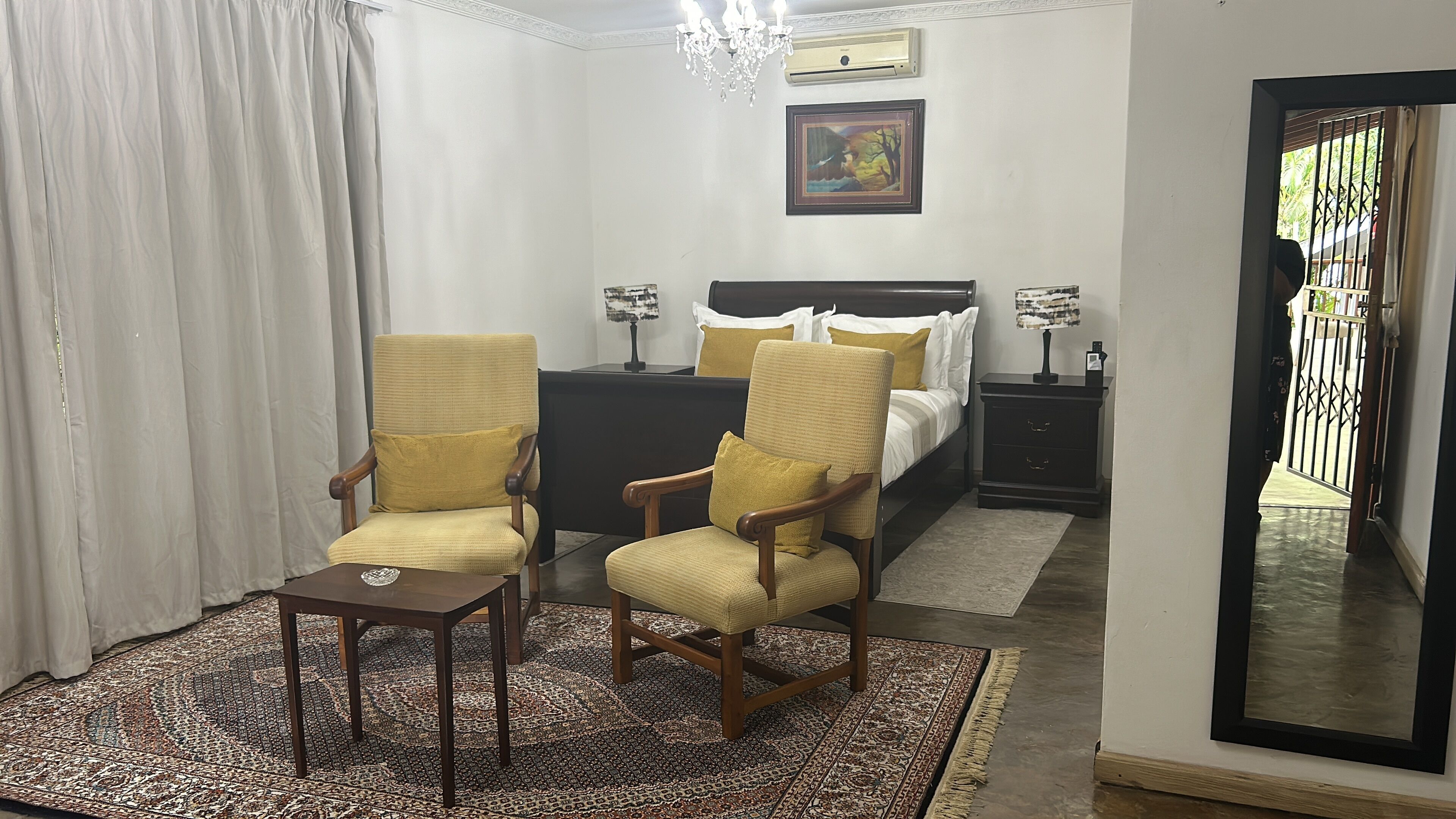 A hotel room with two armchairs, a bed, a nightstand, a mirror, and a patterned rug.