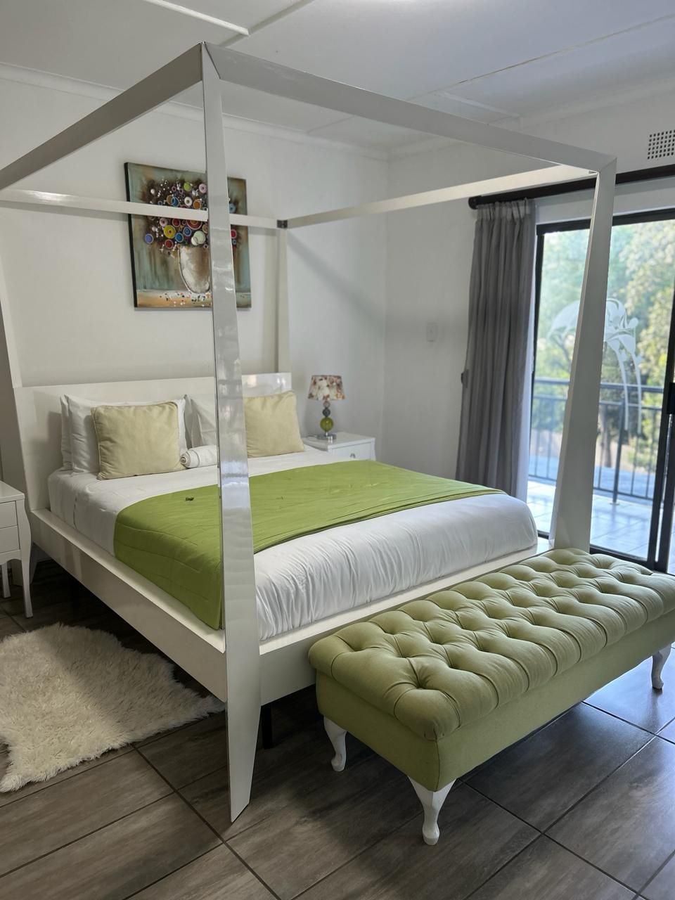 A four-poster bed with a green bedspread and matching bench, a white nightstand with a lamp, and a painting on the wall.