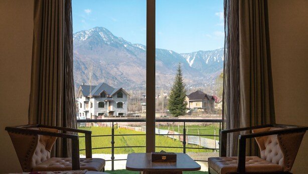 A room with two chairs and a small table, a bed, and a large window offering a view of mountains and houses. - The Sarai (Srinagar)