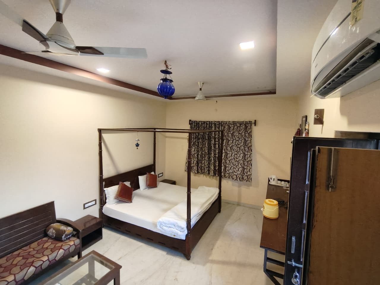 A bedroom with a four-poster bed, a ceiling fan, a wall-mounted air conditioner, and a bedside table with a lamp.