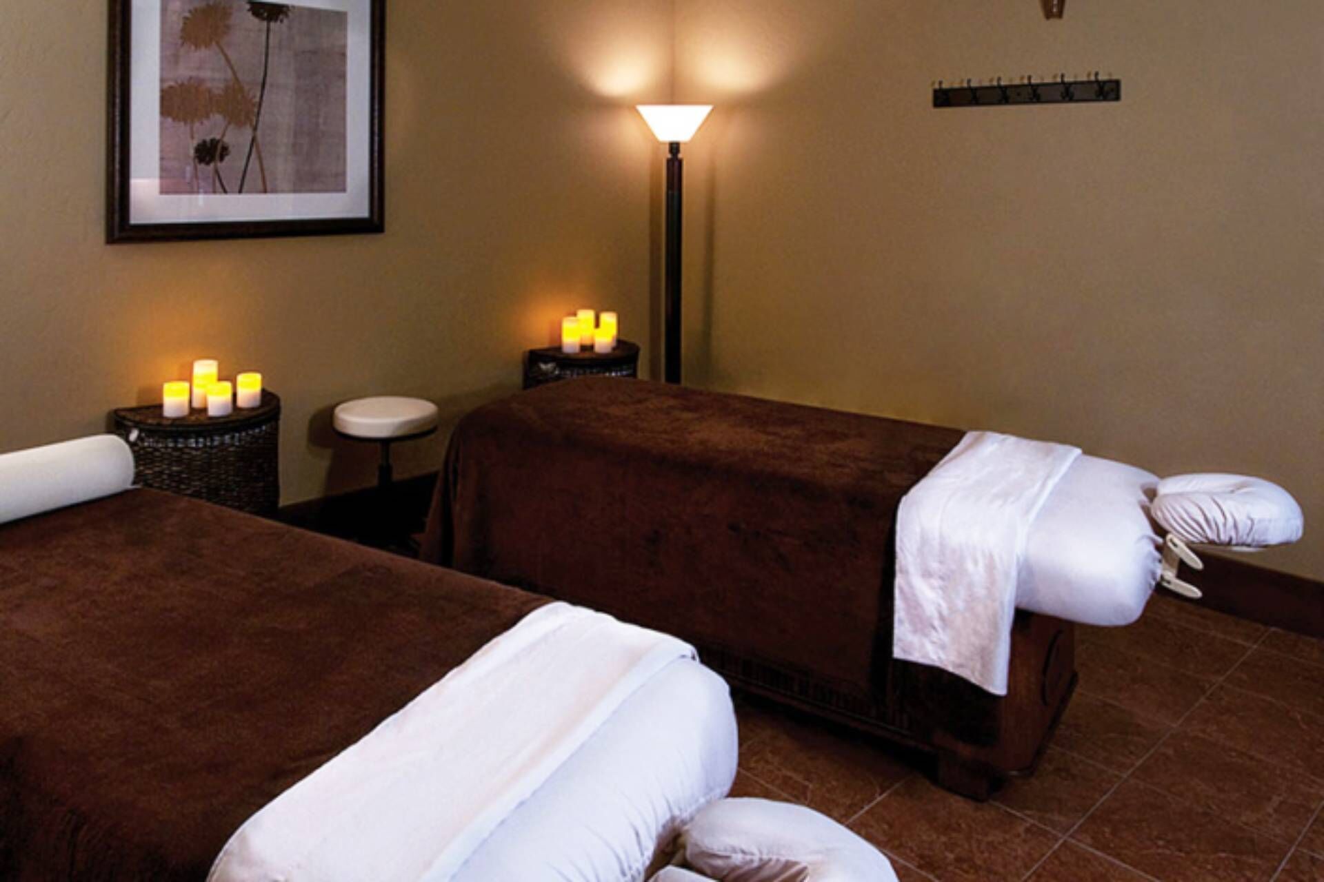 A spa room with two massage tables, a wall-mounted lamp, and candles on a side table.