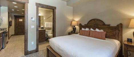 Un dormitorio con una cama grande, cabecera de madera, dos lámparas de noche, una mesita de noche de madera y un baño visible a través de una puerta abierta.