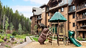 Zona de juegos infantiles con un tobogán y estructuras para escalar, rodeada por un edificio de madera con balcones.