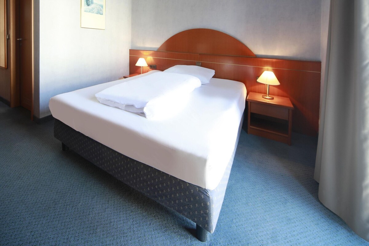 A hotel room with a large bed, two bedside tables with lamps, a wooden headboard, and a carpeted floor.