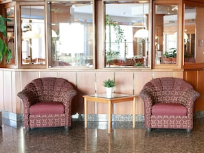 A lobby with two patterned armchairs, a small table with a potted plant, and a large window with a view of outdoor seating.