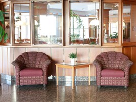 A lobby with two patterned armchairs, a small table with a potted plant, and a large window with a view of outdoor seating.