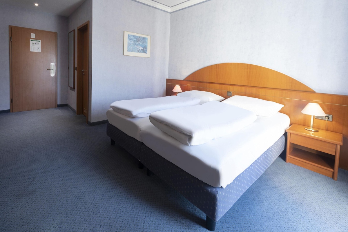 A hotel room with a double bed, two bedside tables with lamps, a wooden headboard, a framed picture on the wall, and a carpeted floor.