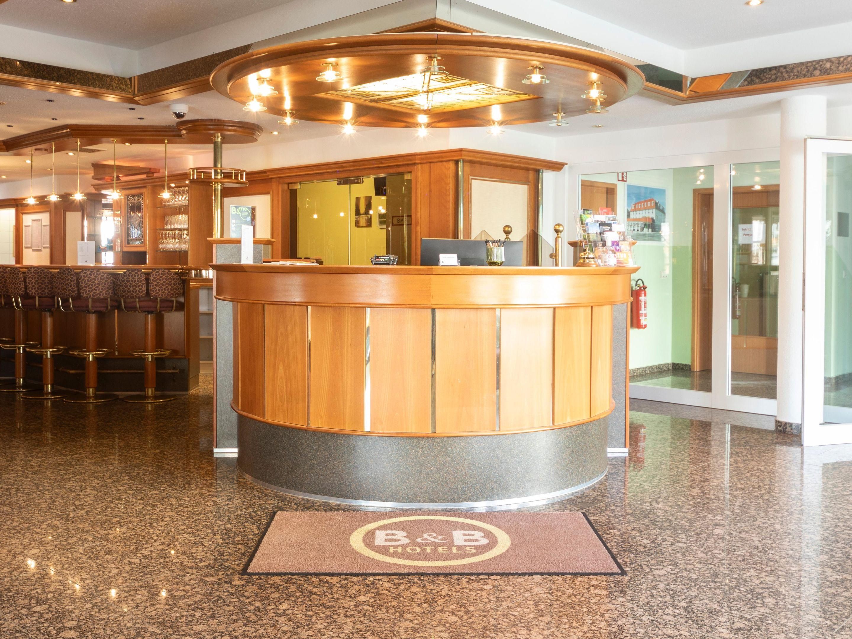 A hotel reception area with a wooden reception desk, a bar with stools, and a glass door entrance.