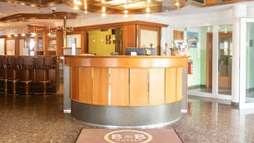 A hotel reception area with a wooden reception desk, a bar with stools, and a glass door entrance.